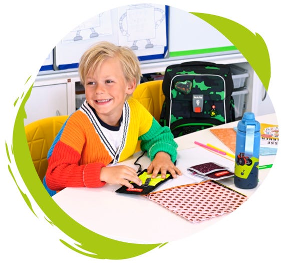 Kind sitzt im Klassenzimmer neben dem Schulranzen und hält einen Brustbeutel in der Hand