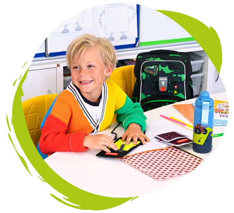 Kind sitzt im Klassenzimmer neben dem Schulranzen und hält einen Brustbeutel in der Hand