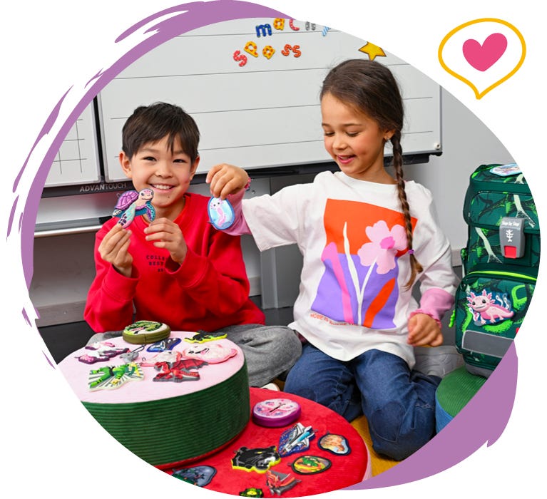 Zwei Kinder pr&auml;sentieren austauschbare Motiv-Applikationen vor einem gr&uuml;nen Schulranzen im Klassenzimmer.