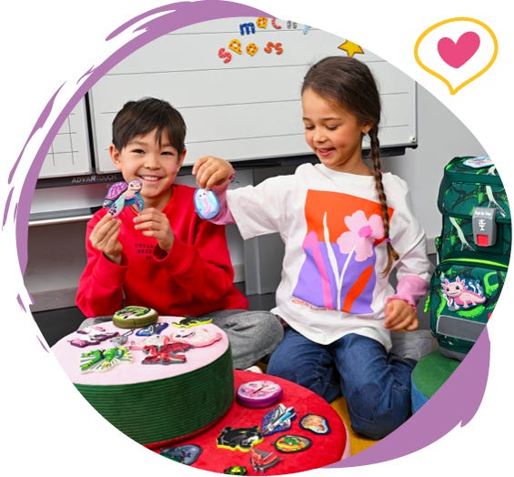 Zwei Kinder pr&auml;sentieren austauschbare Motiv-Applikationen vor einem gr&uuml;nen Schulranzen im Klassenzimmer.