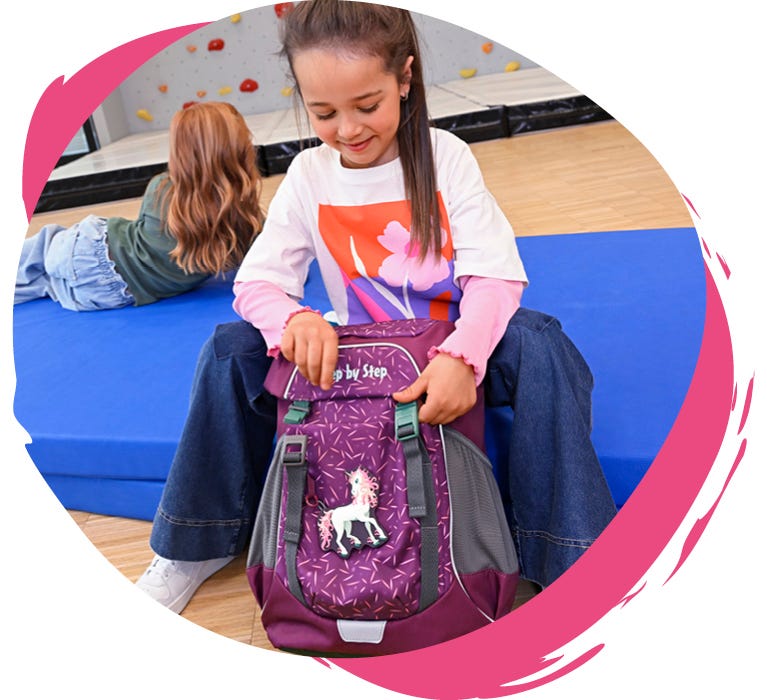 Kind &ouml;ffnet einen violetten Rucksack mit Einhornmotiv in einer Sporthalle mit Kletterwand.
