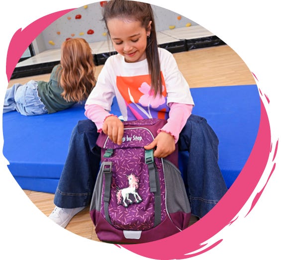 Kind &ouml;ffnet einen violetten Rucksack mit Einhornmotiv in einer Sporthalle mit Kletterwand.