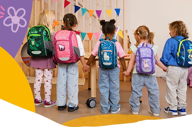 Fünf Kinder mit unterschiedlich gestalteten Kindergartenrucksäcken stehen Hand in Hand in einem Gruppenraum.