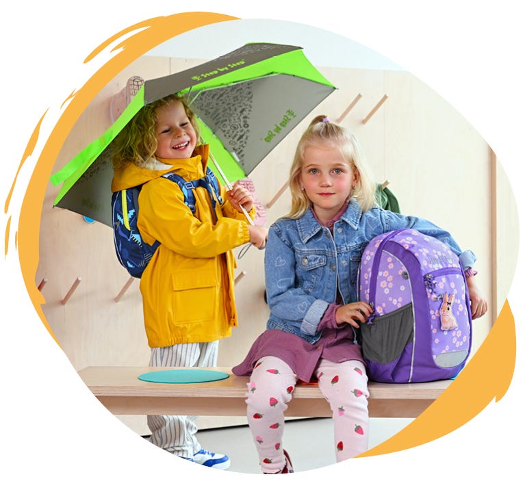 KIGA MAXI Zwei Kinder mit Kindergartenrucksäcken in einer Garderobe, eines mit Regenschirm und Regenjacke, das andere mit violettem Rucksack mit Hasenmotiv.
