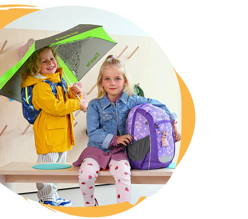 KIGA MAXI Zwei Kinder mit Kindergartenrucksäcken in einer Garderobe, eines mit Regenschirm und Regenjacke, das andere mit violettem Rucksack mit Hasenmotiv.