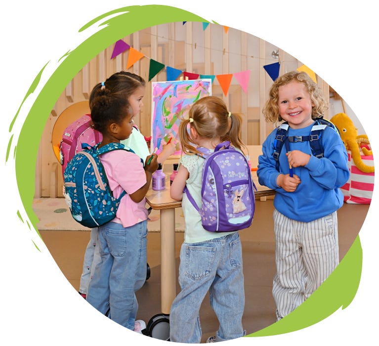 KIGA MINI Vier Kinder mit bunten Kindergartenrucksäcken malen und basteln gemeinsam in einem Gruppenraum.