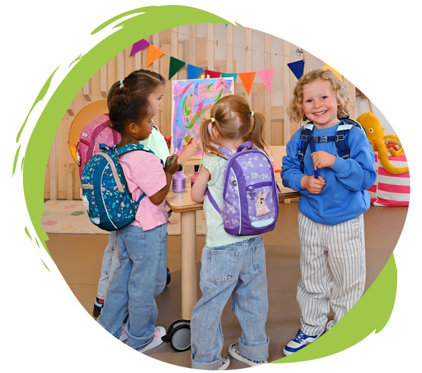 KIGA MINI Vier Kinder mit bunten Kindergartenrucksäcken malen und basteln gemeinsam in einem Gruppenraum.