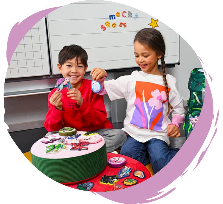 MAGIC MAGS Zwei Kinder präsentieren austauschbare Magic-Mags-Motive vor einem Schulranzen im Klassenzimmer.