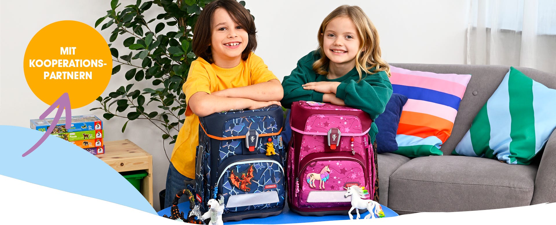 Zwei Kinder pr&auml;sentieren Schulranzen mit Drachen- und Einhornmotiven in einem Wohnraum. Text: &sbquo;Mit Kooperationspartnern&lsquo;
