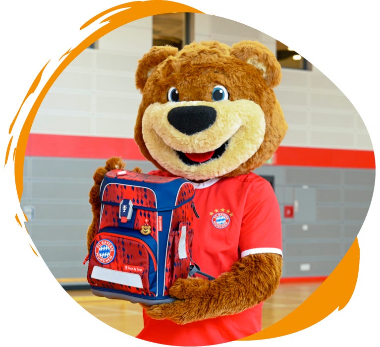 B&auml;ren-Maskottchen pr&auml;sentiert einen rot-blauen Schulranzen im FC-Bayern-M&uuml;nchen-Design in einer Sporthalle