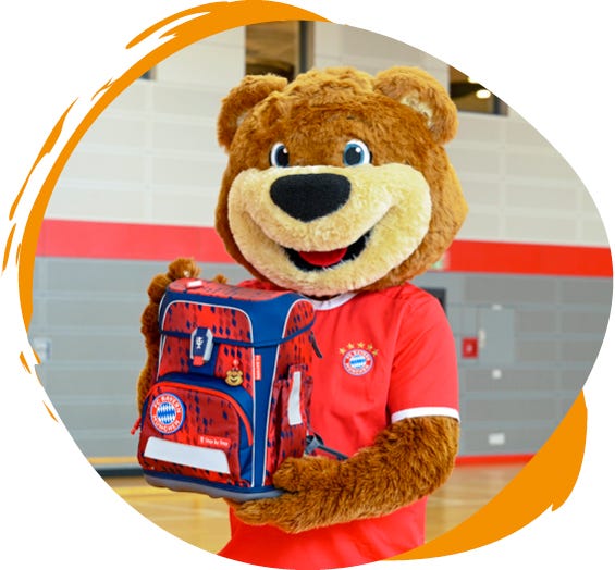 B&auml;ren-Maskottchen pr&auml;sentiert einen rot-blauen Schulranzen im FC-Bayern-M&uuml;nchen-Design in einer Sporthalle