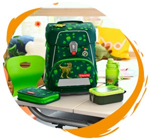 Motivwelten Dunkelgrüner Schulranzen mit Dinosauriermotiv sowie passendes Federmäppchen, Trinkflasche und Brotdose auf einem Schultisch.