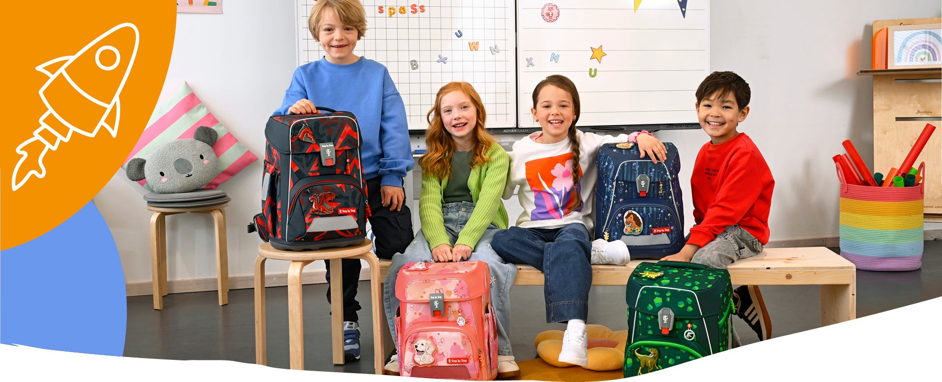 Vier Kinder präsentieren Schulranzen mit Drachen-, Hunde-, Weltraum- und Dinosauriermotiven im Klassenzimmer.