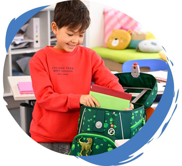 Schulranzen richtig packen Kind packt Schulhefte in einen geöffneten Schulranzen mit Dinosaurier-Motiv im Klassenzimmer.