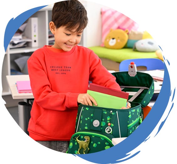 Schulranzen richtig packen Kind packt Schulhefte in einen geöffneten Schulranzen mit Dinosaurier-Motiv im Klassenzimmer.